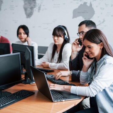 Woman in a denim jacket on a phone at a desk with a laptop; colleagues wearing headsets and working on computers in the background in an office.