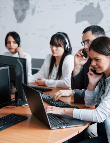 Woman in a denim jacket on a phone at a desk with a laptop; colleagues wearing headsets and working on computers in the background in an office.