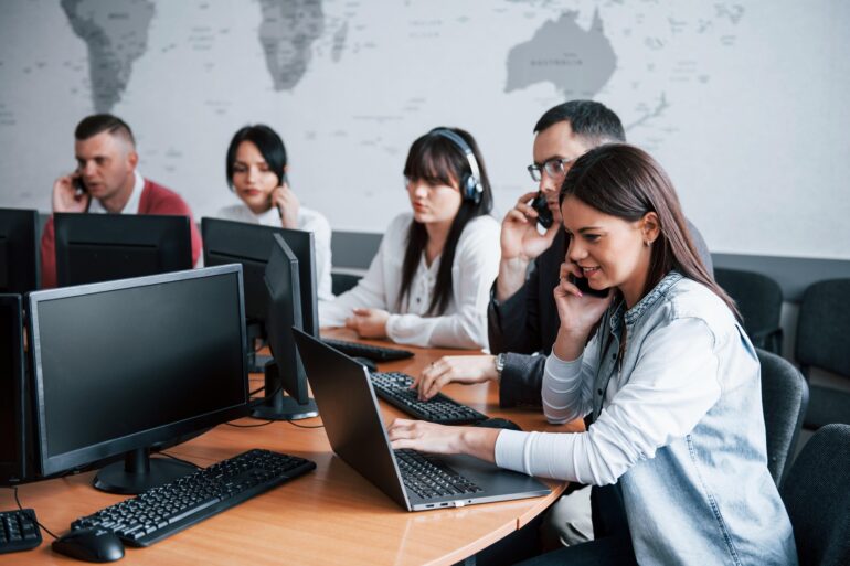 Woman in a denim jacket on a phone at a desk with a laptop; colleagues wearing headsets and working on computers in the background in an office.