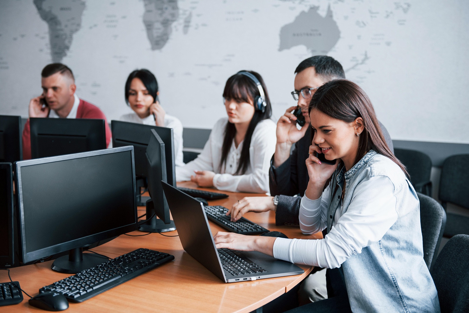 Woman in a denim jacket on a phone at a desk with a laptop; colleagues wearing headsets and working on computers in the background in an office.