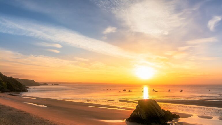 Sunset over a calm sea with a rocky foreground and several boats on the horizon