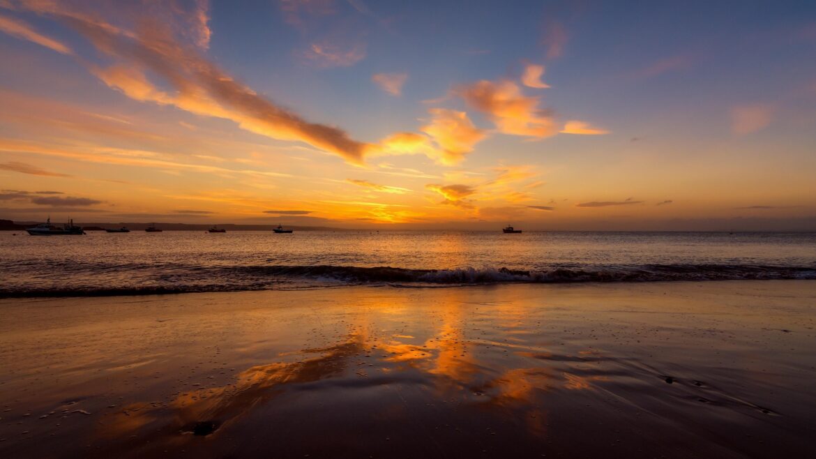 Sunset over calm sea with an orange sky and several boats near the horizon on the water.