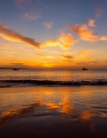 Sunset over calm sea with an orange sky and several boats near the horizon on the water.