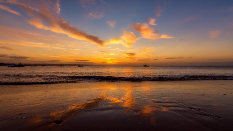Sunset over calm sea with an orange sky and several boats near the horizon on the water.