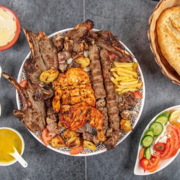 Large platter of grilled meats (ribs and kebabs) with fries and tomatoes, surrounded by sauces and a basket of flatbread