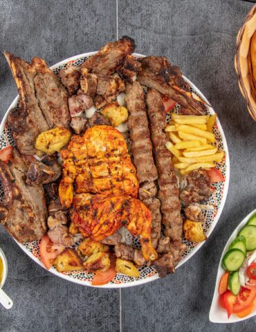Large platter of grilled meats (ribs and kebabs) with fries and tomatoes, surrounded by sauces and a basket of flatbread
