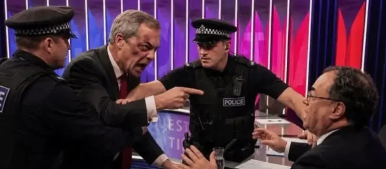 Politician yells at a man seated at a desk as two police officers separate them on a TV set.