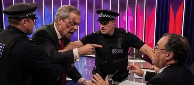Politician yells at a man seated at a desk as two police officers separate them on a TV set.