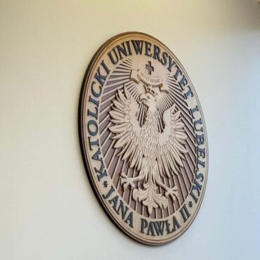 Circular wooden plaque on a pale wall, featuring a detailed eagle emblem with Polish text around the edge (seal of a university).