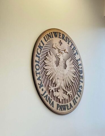 Circular wooden plaque on a pale wall, featuring a detailed eagle emblem with Polish text around the edge (seal of a university).
