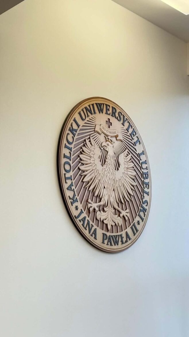 Circular wooden plaque on a pale wall, featuring a detailed eagle emblem with Polish text around the edge (seal of a university).