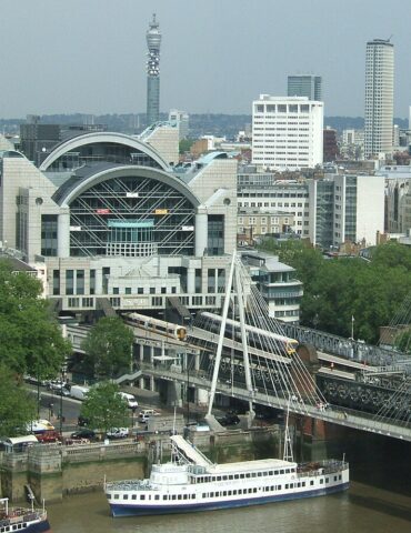 City waterfront with a modern arched train station, a white cable-stayed footbridge, and a riverboat on the river edge.