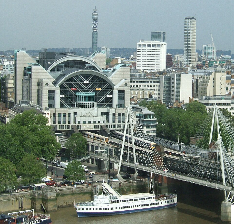 City waterfront with a modern arched train station, a white cable-stayed footbridge, and a riverboat on the river edge.