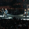 Rock band performing on circular stage under spotlights with a large cheering audience in a crowded arena at a concert.