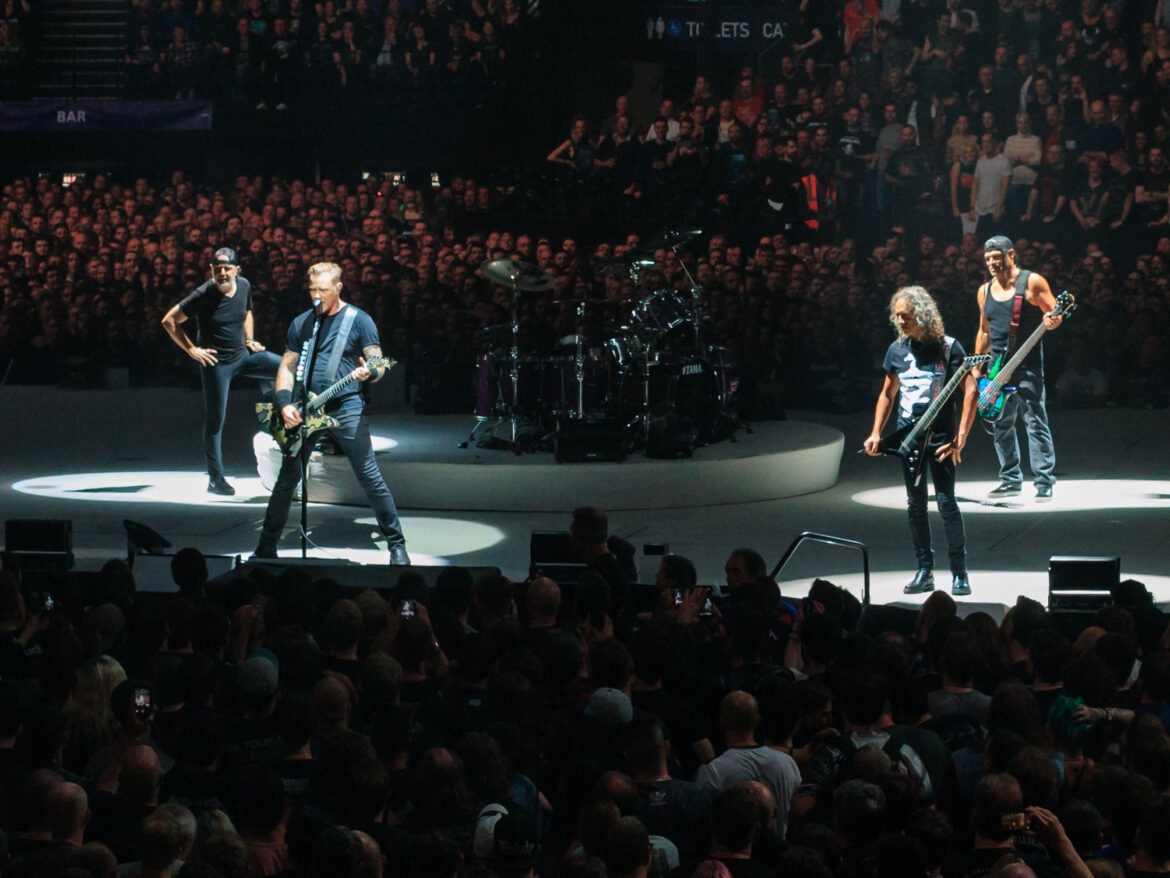 Rock band performing on circular stage under spotlights with a large cheering audience in a crowded arena at a concert.