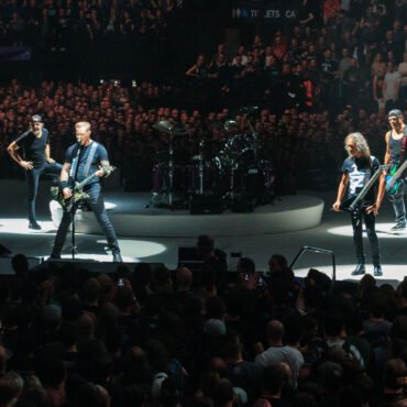 Rock band performing on circular stage under spotlights with a large cheering audience in a crowded arena at a concert.