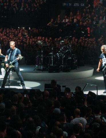 Rock band performing on circular stage under spotlights with a large cheering audience in a crowded arena at a concert.