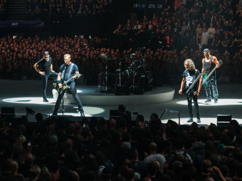 Rock band performing on circular stage under spotlights with a large cheering audience in a crowded arena at a concert.