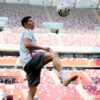 Soccer player in a light gray shirt and shorts juggling a ball in a red-and-white stadium stands behind him