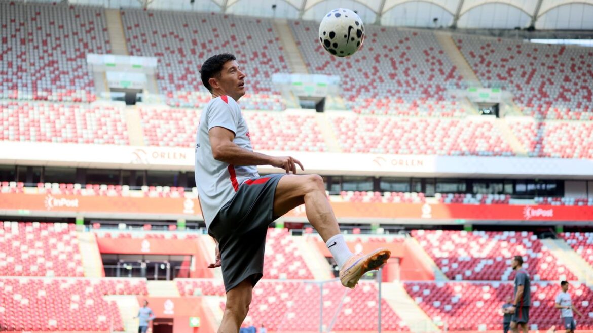 Soccer player in a light gray shirt and shorts juggling a ball in a red-and-white stadium stands behind him