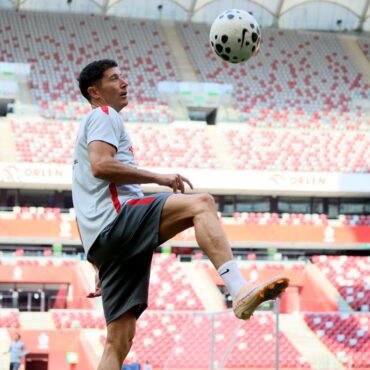 Soccer player in a light gray shirt and shorts juggling a ball in a red-and-white stadium stands behind him