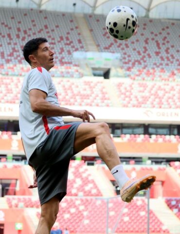 Soccer player in a light gray shirt and shorts juggling a ball in a red-and-white stadium stands behind him