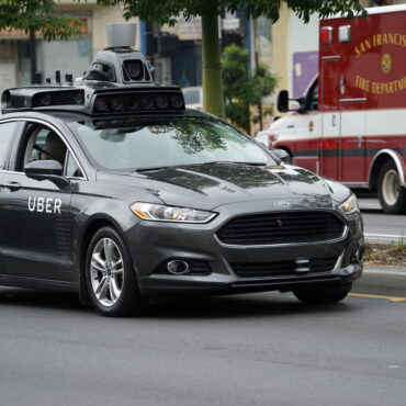 Autonomous Uber vehicle with a roof-mounted sensor rig driving on a city street, with a San Francisco Fire Department ambulance in the background.