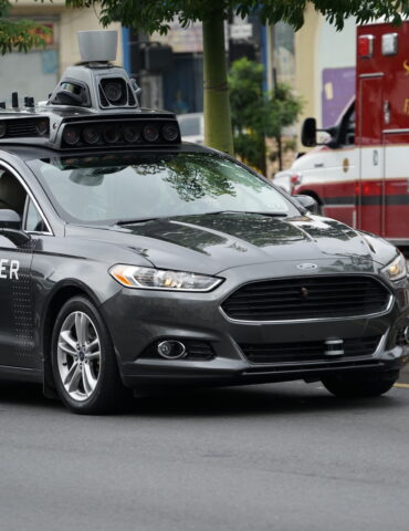 Autonomous Uber vehicle with a roof-mounted sensor rig driving on a city street, with a San Francisco Fire Department ambulance in the background.