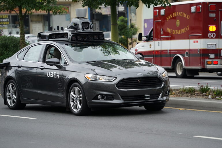Autonomous Uber vehicle with a roof-mounted sensor rig driving on a city street, with a San Francisco Fire Department ambulance in the background.