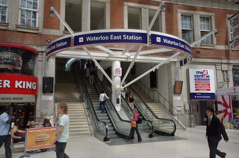 Entrance to Waterloo East Station with escalators and a circular blue sign overhead reading 'Waterloo East Station' and surrounding buildings. A Burger King on the left and a medical centre on the right are visible. People are walking and using the escalators.