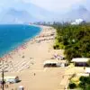Busy sandy beach along a blue sea with numerous colorful umbrellas and sunbathers; mountains and city skyline in the distance.