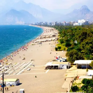 Busy sandy beach along a blue sea with numerous colorful umbrellas and sunbathers; mountains and city skyline in the distance.