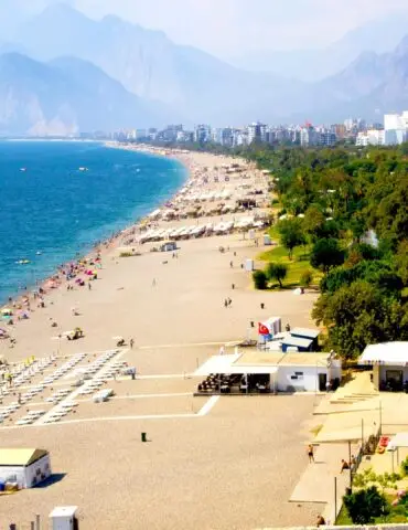 Busy sandy beach along a blue sea with numerous colorful umbrellas and sunbathers; mountains and city skyline in the distance.