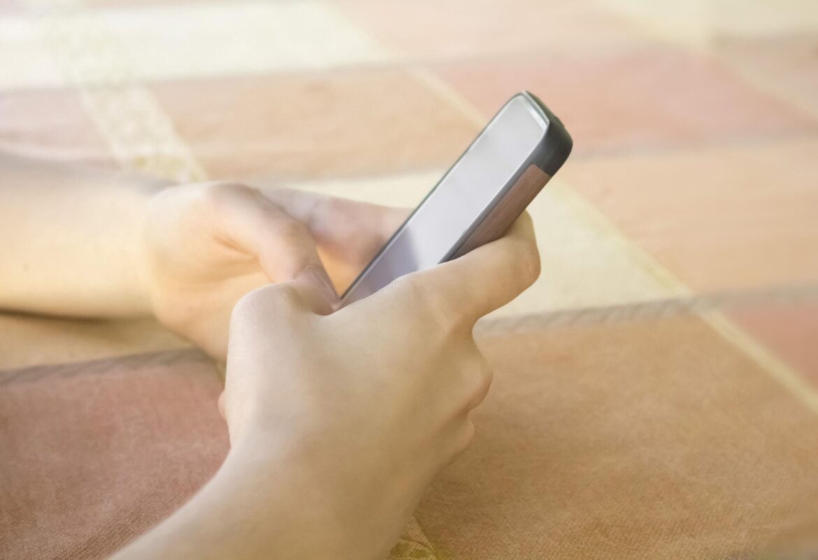 Person holding a smartphone in bed, screen blank, close-up on hands and device against peach-colored blanket