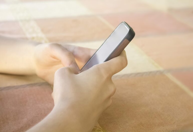 Person holding a smartphone in bed, screen blank, close-up on hands and device against peach-colored blanket
