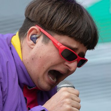 Close-up of a performer singing into a handheld microphone, wearing red sunglasses and a purple jacket with a yellow lining visible.
