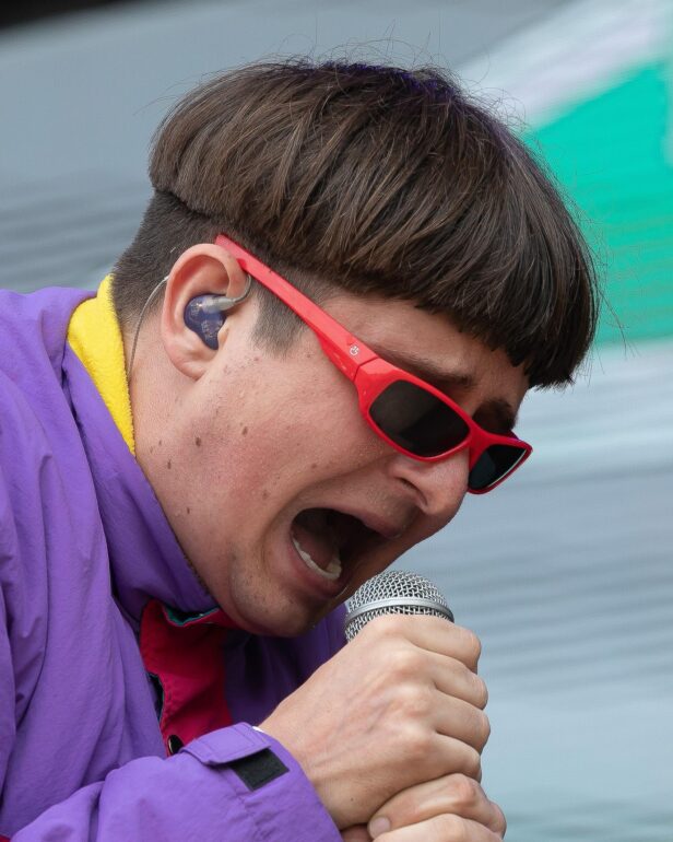 Close-up of a performer singing into a handheld microphone, wearing red sunglasses and a purple jacket with a yellow lining visible.