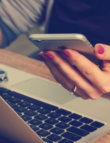Person typing on a smartphone held over a laptop on a wooden desk, with a car key fob nearby.