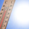 Wooden outdoor thermometer leaning against a bright blue sky with the sun, showing temperatures up to 50 (both scales visible).