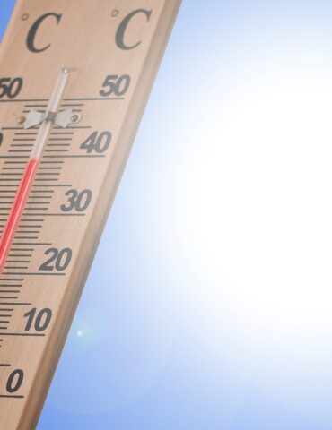Wooden thermometer angled across a clear blue sky with bright sun in the background; red liquid shows a warm temperature.