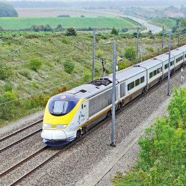 High-speed electric passenger train on elevated tracks through green countryside with overhead wires visible in the landscape