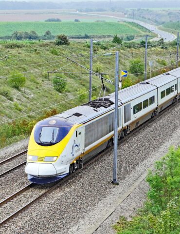 High-speed electric passenger train on elevated tracks through green countryside with overhead wires visible in the landscape