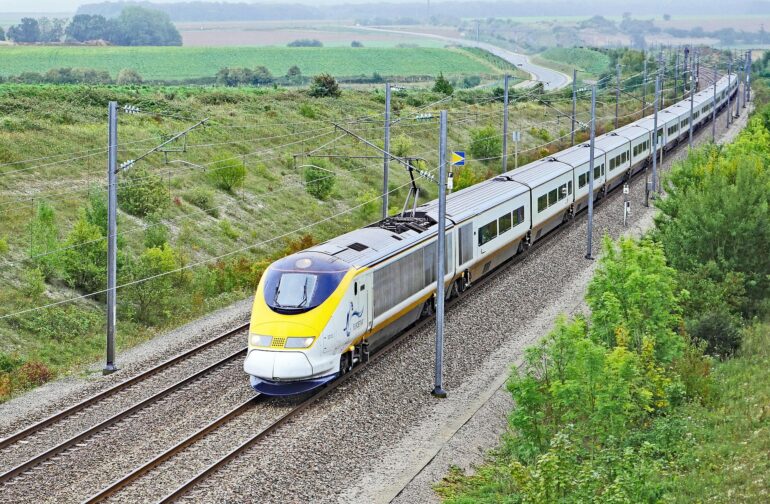 High-speed electric passenger train on elevated tracks through green countryside with overhead wires visible in the landscape