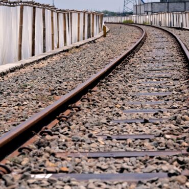 Railroad tracks curve to the right beside a gravel bed and a long white plastic fence on the left.