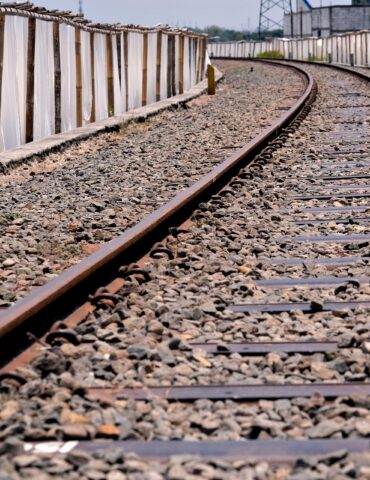Railroad tracks curve to the right beside a gravel bed and a long white plastic fence on the left.