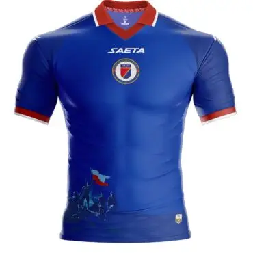 Blue Haiti national football jersey with red collar and white trim, features crest and SAETA logo on chest and stylized silhouettes at bottom left