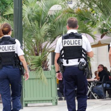 Three police officers in tactical vests walking away in an outdoor plaza with palm trees and diners in the background, seen from behind.