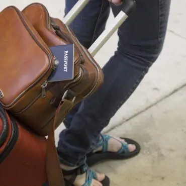 Traveler gripping a suitcase handle with a brown leather backpack; a passport peeks from the backpack pocket.