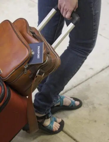 Traveler gripping a suitcase handle with a brown leather backpack; a passport peeks from the backpack pocket.