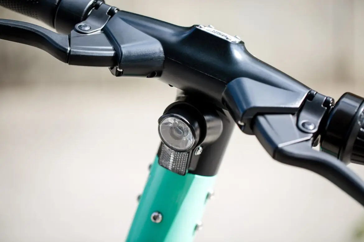 Close-up of a black bicycle handlebar stem with a circular front light mounted on the teal frame area beneath.
